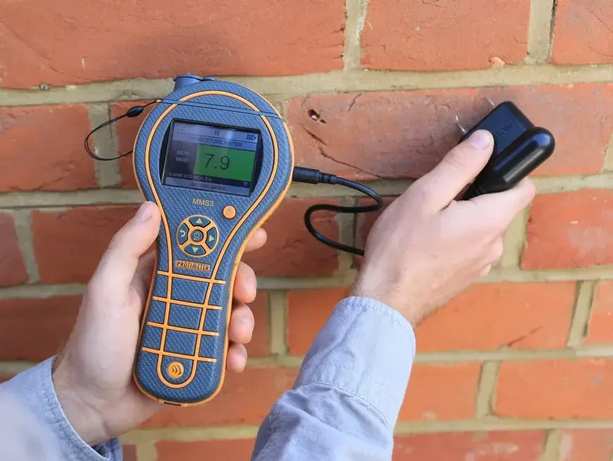 A moisture meter pressed against a brick wall to check how far water soaked in after a leak