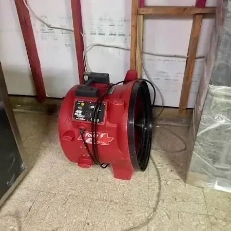A red air mover running beside a wall opened at the bottom, with plastic sheeting hung to contain the work area
